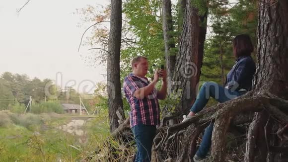 男子在针叶林用手机拍摄女子快乐的女人在松林的树根上摆姿势移动视频的预览图