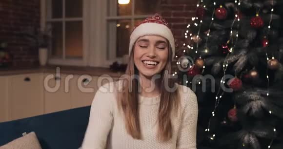 戴着圣诞帽的年轻漂亮女孩微笑着坐在圣诞树旁挥舞着手拿着红色礼物盒视频的预览图