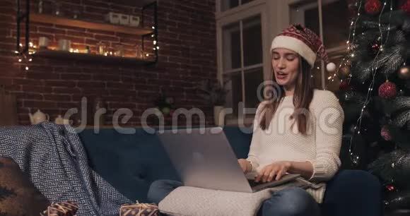 快乐的白种人女孩戴着圣诞帽坐在沙发上在科西家背景靠近圣诞树妇女视频的预览图