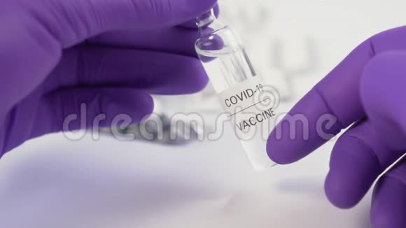 手拿冠状病毒疫苗安瓿戴蓝色防护手套白色桌子上有模糊的药丸视频的预览图