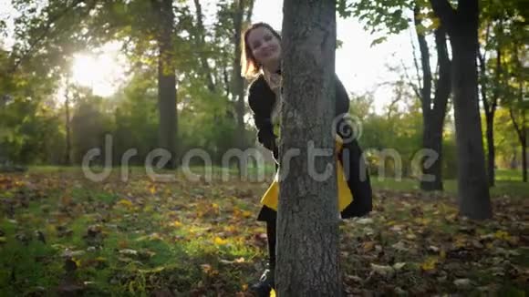 和妈妈一起玩一个迷人的微笑女孩和她的父母一起在秋天的公园里玩一家人躲在树干后面视频的预览图