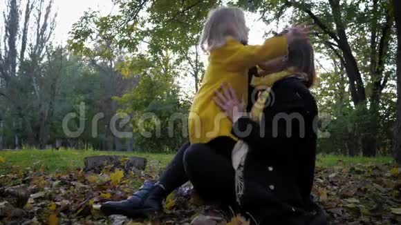 放松年轻的母亲和她心爱的女儿喜欢在秋天的公园里放松坐在满是树叶的空地上视频的预览图