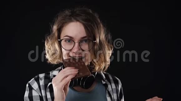 将带眼镜和耳机的年轻女性靠近假扮成玩吃的样子嚼着巧克力棒和坚果视频的预览图