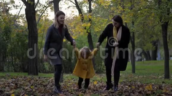 两个美丽的年轻女子和微笑的小女儿在秋天的草地上玩耍她的手在草地上撒满了树叶视频的预览图