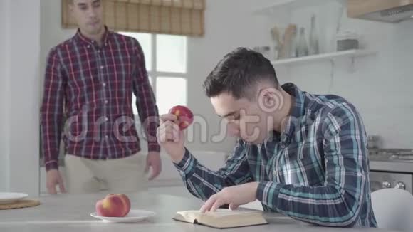 成年白种人黑发男子坐在桌子旁看书吃红苹果他的孪生兄弟走了过来视频的预览图