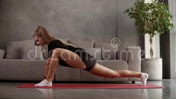 头发长长的金发碧眼的女人穿着黑色运动服在家在灰色沙发前的垫子上训练伸展肌肉视频的预览图
