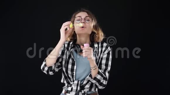 戴眼镜的快乐女孩的肖像在黑色背景上用棍子吹了很多肥皂泡演播室的录像视频的预览图