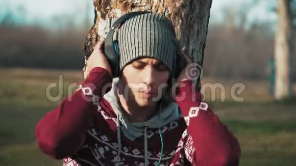 戴帽子的年轻白种人听着耳机里的音乐唱歌在森林模糊的背景上特写视频的预览图