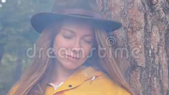 带着黄色雨衣和帽子的红发旅游女孩的画像透过火烟看着镜头视频的预览图