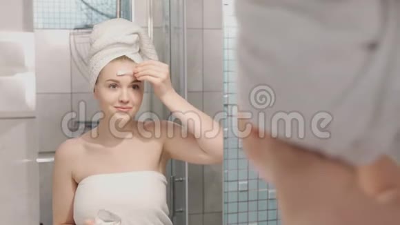 一张美丽的年轻女子的肖像穿着浴袍和毛巾站在浴室里在浴室下面敲击补片视频的预览图