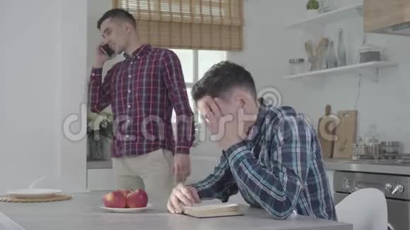 愤怒的白种人坐在桌子旁揉着太阳穴他的孪生兄弟在智能手机上聊天视频的预览图