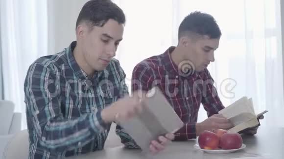 成年白种人黑发男子阅读他的孪生兄弟在平板电脑上玩游戏把书收起来看着视频的预览图