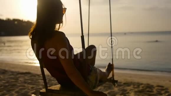 在夕阳的照耀下快乐的女人在海边的秋千上年轻女子度假时正在海滩上放松视频的预览图