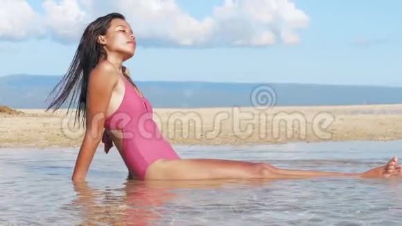 迷人的亚洲女孩日光浴坐在海水中沙滩侧视视频的预览图