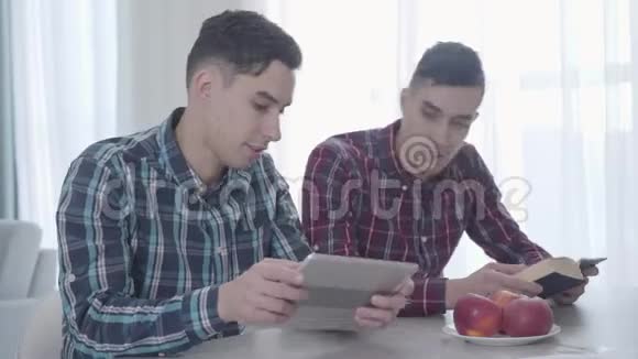 成年白种人在平板电脑上玩电子游戏的肖像并展示给他的孪生兄弟布鲁内特看书视频的预览图