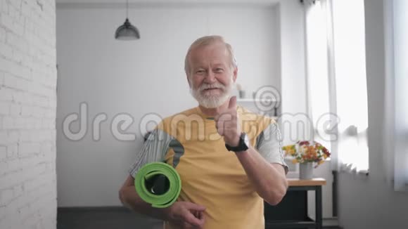 健康护理英俊的微笑老人身材魁梧手里拿着瑜伽垫运动后拿着一只智能手表视频的预览图
