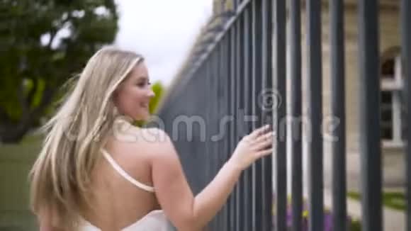 一个夏天年轻漂亮的金发女郎穿着白嫩嫩的裙子走在格子铁栅栏附近行动后视图视频的预览图
