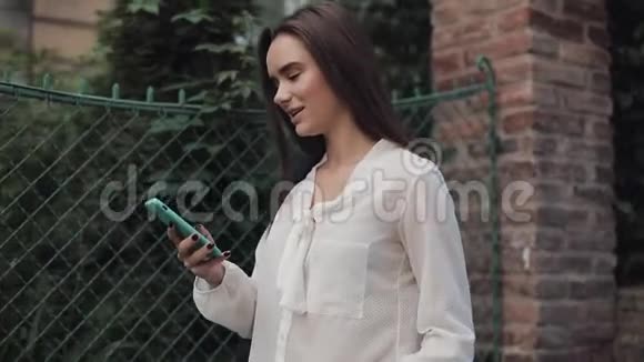 漂亮的学生女孩一边走在舒适的老城街一边用她的电话一边微笑着环顾四周视频的预览图