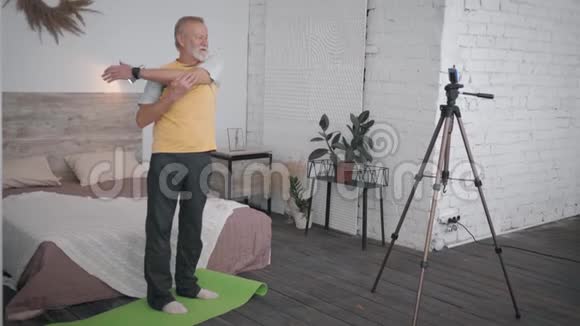 迷人的老人在舒适的房间里为社交网络和追随者录制视频智能手机展示健康锻炼视频的预览图