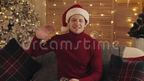穿着圣诞帽和冬季毛衣坐在沙发上抱着礼物挥手致意的年轻人的肖像视频的预览图