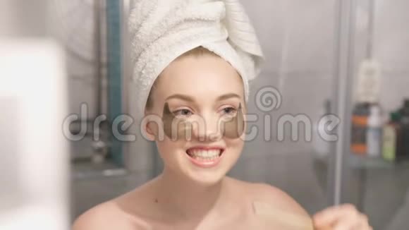 一张美丽的年轻女子的肖像穿着浴袍和毛巾站在浴室里在浴室下面敲击补片视频的预览图
