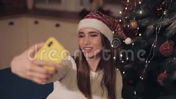 快乐的年轻漂亮女孩戴着圣诞帽坐在圣诞树旁的沙发上自拍挥手视频的预览图