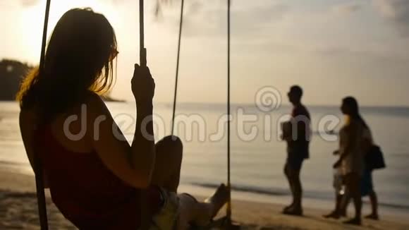 在夕阳的照耀下快乐的女人在海边的秋千上年轻女子度假时正在海滩上放松视频的预览图