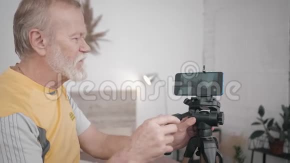 现代祖父一位年长的男性博客作者在智能手机上安装了摄像头在他的房间里为vlog创建视频内容视频的预览图