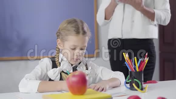 白种人女孩的肖像扎着辫子写在练习本上作为成年私人教师站在她旁边儿童视频的预览图
