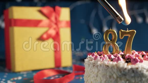 白色生日蛋糕87号金色蜡烛用打火机点燃蓝色背景灯和礼品黄色盒子用红色系着视频的预览图