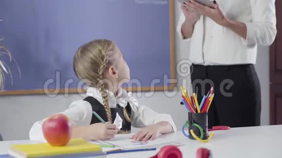聪明的白种人女孩扎着辫子回头看着无法辨认的导师写在练习本上学校教育视频的预览图