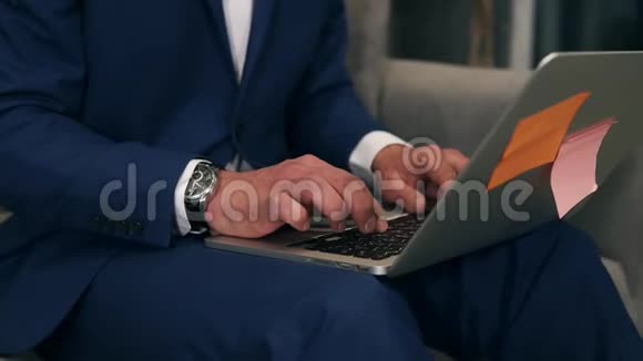 商人手在笔记本电脑键盘上打字而坐在沙发上膝盖上抱着灰色的笔记本电脑一个男人视频的预览图
