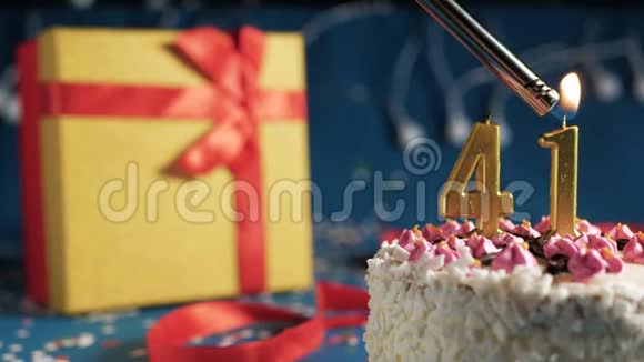 白色生日蛋糕编号41金蜡烛用打火机点燃蓝色背景灯和礼品黄色盒子系好视频的预览图