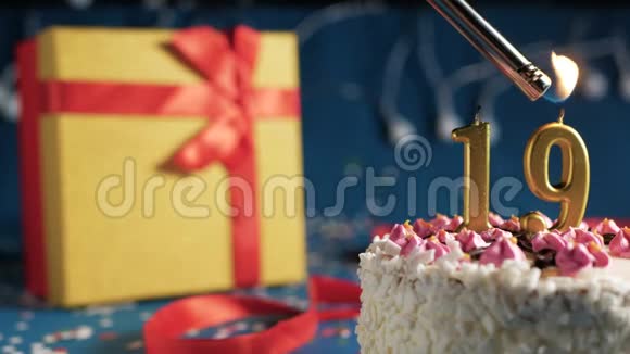 白色生日蛋糕19号金色蜡烛由打火机燃烧蓝色背景灯和礼品黄色盒子绑在一起视频的预览图