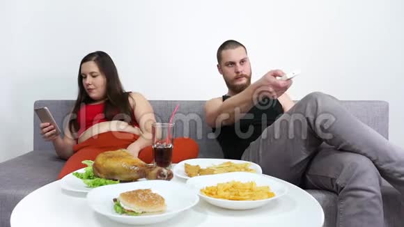 超重夫妇吃快餐汉堡包薯条炸薯条和看电视视频的预览图