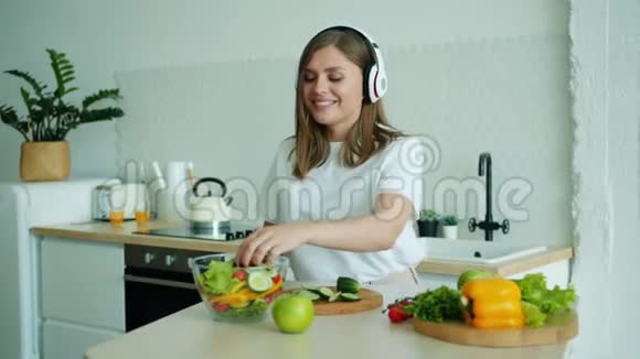 戴着耳机的快乐女人在家里做沙拉跳舞听音乐视频的预览图