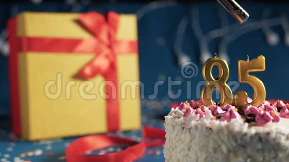 白色生日蛋糕85根金色蜡烛用打火机点燃蓝色背景黄色礼品盒装有红色视频的预览图