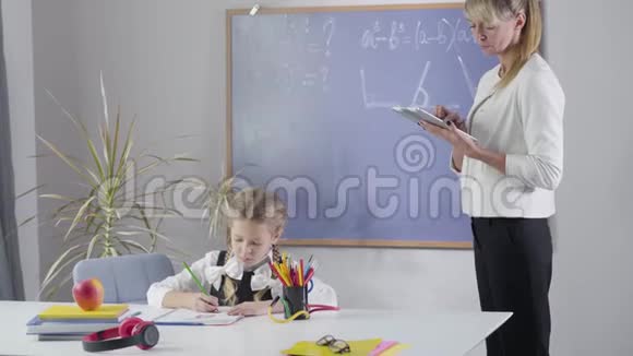 一个留着辫子的高加索小女孩在练习本上作为成人私人教师在平板电脑上搜索任务儿童视频的预览图