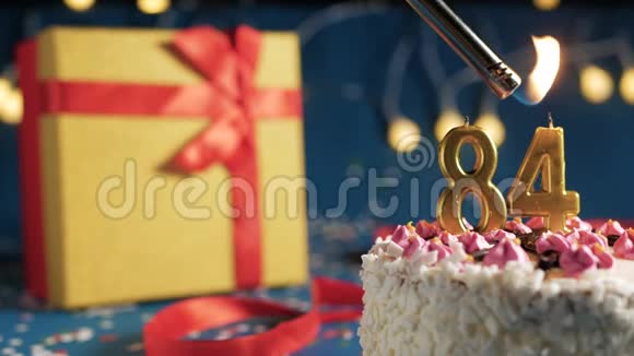 白色生日蛋糕84根金色蜡烛用打火机点燃蓝色背景黄色礼品盒装有红色视频的预览图