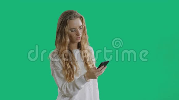 年轻漂亮的女士戴着时髦的眼镜手里拿着一部智能手机在绿色屏幕上使用智能手机视频的预览图