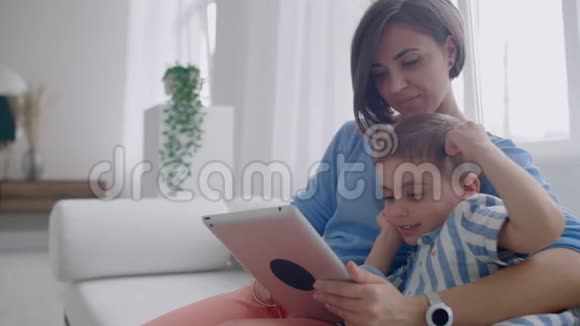 母亲和儿子坐在沙发上使用数字平板电脑快乐的妈妈和小男孩一起使用带有触摸屏的平板电脑视频的预览图