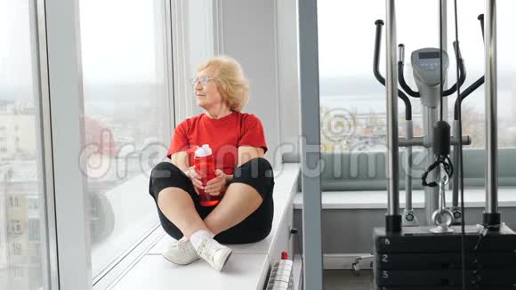 相当活跃的退休妇女穿着红色t恤坐在健身房的窗台上老年人拿着水瓶和水冷静视频的预览图