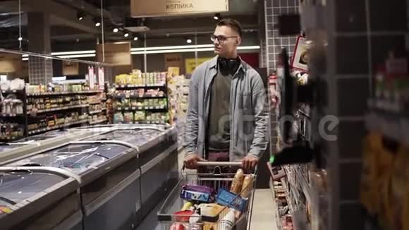 在当地超市购物时戴着眼镜戴着耳机推着手推车装满食物的快乐男人视频的预览图