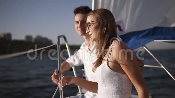 一对年轻夫妇在游艇上共度时光的侧视图一个穿着白裙子戴着太阳镜坐在一起的女孩视频的预览图