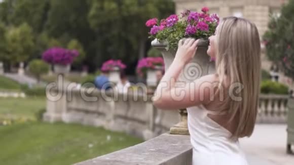 穿着白色性感礼服的金发碧眼在夏日的绿色公园里抚摸着她的长发背景是紫色的花坛视频的预览图