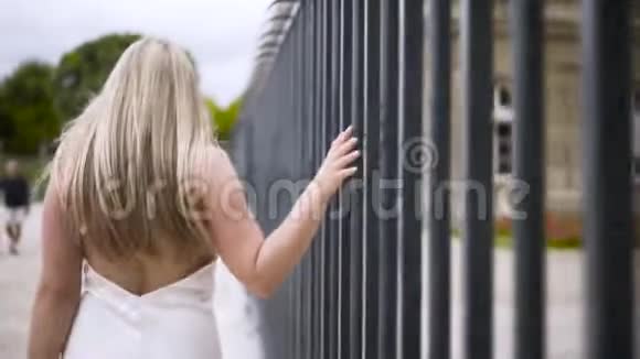 一个年轻的女人穿着白色性感的裙子赤裸的背部沿着铁栅栏走着摸着它行动视频的预览图
