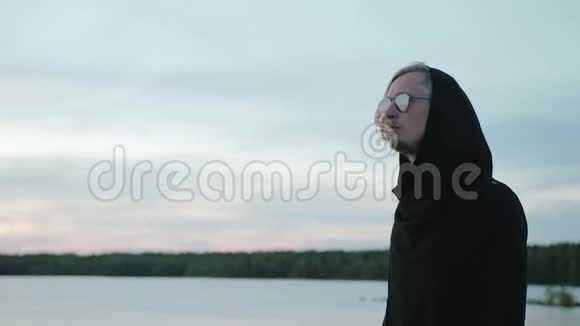 一个英俊的男人的肖像一个穿着黑色引擎盖的金发沿着一个美丽的大湖散步旅行理念独特视频的预览图