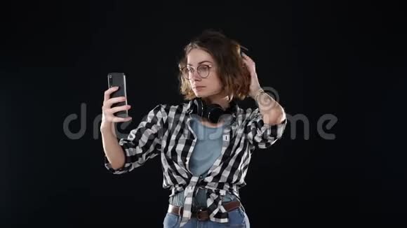 可爱的年轻女子穿着格子衬衫脖子上戴着耳机边看着自己的智能手机一边自拍视频的预览图