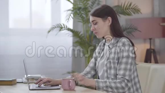 年轻的白种人黑发女人在桌子旁放着茶或咖啡在笔记本电脑键盘上打字视频的预览图