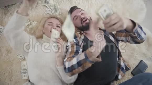 高瞻远瞩的富有高加索夫妇躺在柔软的地毯上分散金钱快乐的年轻男女视频的预览图